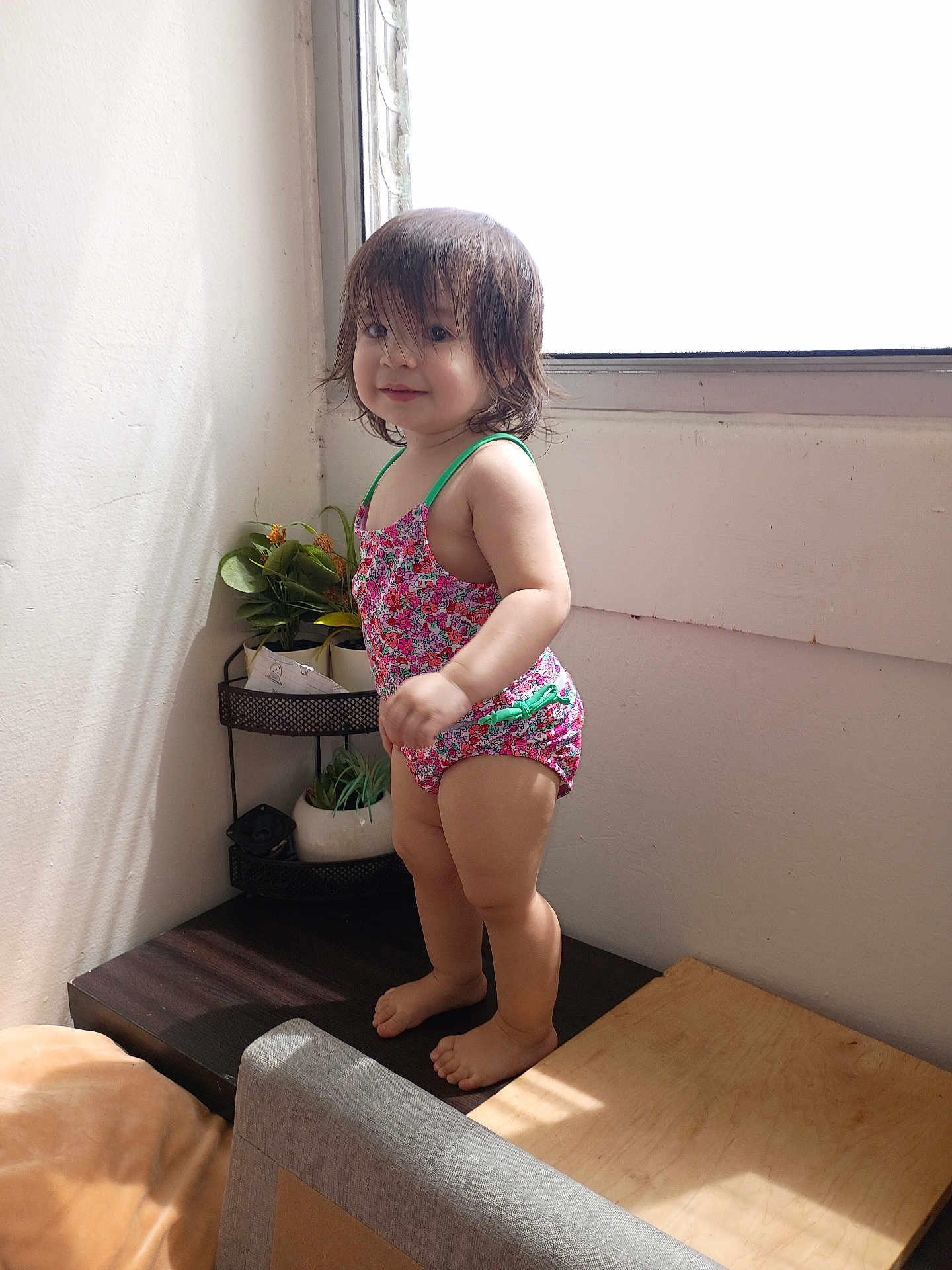 Xion is registered to the contest to win money with this photo: toddler, child, swimsuit, standing, indoor, window, sunlight, plant, greenery, wooden_surface, barefoot, hair, face, smile, corner, shadow, bright, colorful, room, casual