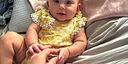 Athena is registered to the contest to win money with this photo: baby, bed, blanket, child, comfort, cute, eyes, face, hand, holding_hands, indoor, infant, person, pillow, playful, sitting, skin, smile, toy, yellow_clothing