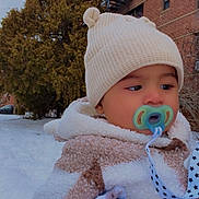 Fernando is registered to the contest to win money with this photo: baby, pacifier, winter_clothing, knit_hat, blanket, snow, outdoor, building, brick_wall, tree, child, cold_weather, face, cute, bundle, warm, portrait, infant, accessory, scarf