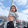 baby, child, hoodie, gray_clothing, outdoor, sky, clouds, hand, person, car, parking_lot, winter_clothing, cute, infant, portrait, casual, daytime, holding, expression, young