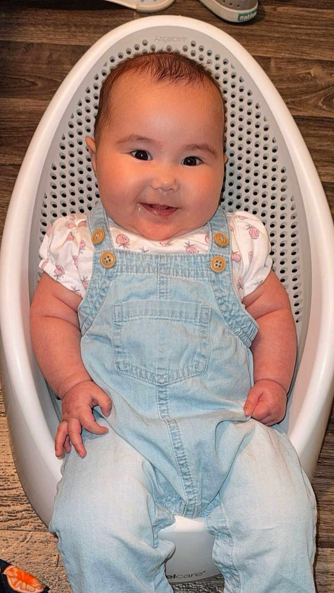 Lena-Sage is registered to the contest to win money with this photo: baby, infant, smiling, drool, denim_overalls, white_shirt, baby_bath_seat, wooden_floor, footwear, face, child, cute, chubby_cheeks, sitting, indoor, clothing, person, happy, baby_toy, casual