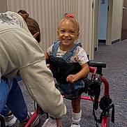 Jazmynn is registered to the contest to win money with this photo: child, toddler, walker, mobility_aid, clinic, hallway, carpet, smile, denim_overalls, pink_bow, shoes, socks, caregiver, assistance, framed_photo, wall, happy, medical, support_device, outfit