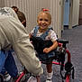 child, toddler, walker, mobility_aid, clinic, hallway, carpet, smile, denim_overalls, pink_bow, shoes, socks, caregiver, assistance, framed_photo, wall, happy, medical, support_device, outfit