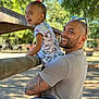 child, toddler, adult, man, smile, laughing, outdoors, park, trees, wooden_rail, sunglasses, tattoo, holding, portrait, candid, hello_kitty, short_sleeves, hair_bun, happy, family_moment
