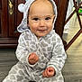 baby, child, onesie, giraffe_pattern, hood, fuzzy_clothing, sitting, floor, wooden_cabinet, smile, cheeky_expression, indoor, furniture, hand, face, person, cute, infant, footwear, hooded_outfit