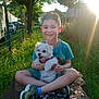 child, boy, dog, pet, grass, tree, sunlight, rock, flower_petals, outdoor, smiling, happy, green_shoes, tshirt, shorts, fence, nature, summer, park, sunshine