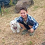 child, boy, dog, pet, grass, rock, fence, car, outdoor, crouching, casual_clothing, sneakers, smiling, happy, animal, mammal, friendly, daylight, nature, playful