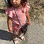 child, girl, curly_hair, pink_top, floral_shorts, white_sandals, black_purse, outdoor, gravel, sunlight, shadow, bracelet, standing, serious_expression, accessory, summer_clothing, nature, daylight, person, casual