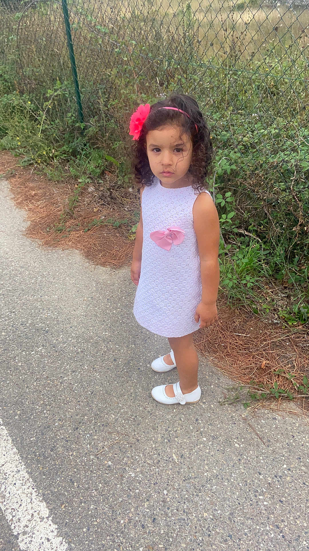 Raissa a rejoint le concours — aidez-le/la à gagner de superbes lots ! child, girl, white_dress, pink_bow, flower_headband, curly_hair, white_shoes, sidewalk, chain_link_fence, greenery, outdoor, nature, serious_expression, standing, person, fashion, cute, young, portrait, casual