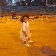 Raissa participe au concours pour gagner de l'argent avec cette photo : child, girl, white_dress, curly_hair, pavement, evening, park, bench, trees, city, buildings, outdoor, sidewalk, shadow, street_lamp, grass, casual, young, standing, curious