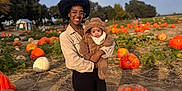 Karter joined the competition — help win amazing prizes! afro_hairstyle, autumn, baby, baby_clothing, black_hair, cloudy_sky, daylight, field, fuzzy_baby_outfit, glasses, holding_baby, leggings, nature, outdoor, pumpkin, pumpkin_patch, smiling, sneakers, trees, woman