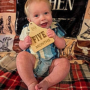 Kohen is registered to the contest to win money with this photo: baby, infant, child, smiling, holding_object, wooden_sign, five_months, blue_eyes, blonde_hair, plaid_blanket, western_theme, cowboy, sitting, cute, portrait, clothing, shorts, happy, indoor, milestone