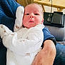 baby, infant, onesie, white_clothing, cute, portrait, person, indoor, adult_arm, holding, blue_sleeve, face, expression, pout, glasses, tablet, wooden_table, home, closeup, newborn