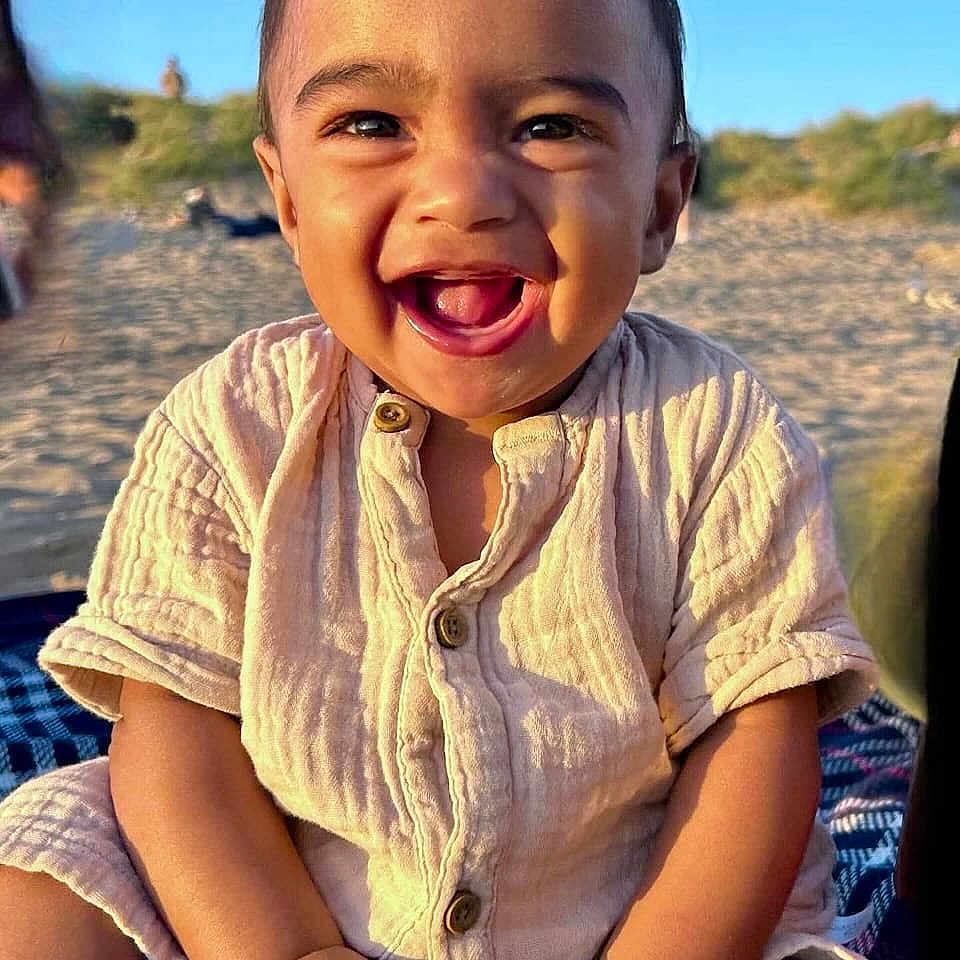 Kenzo is registered to the contest to win money with this photo: baby, beach, blanket, buttoned_shirt, casual_clothing, child, cute, daytime, face, happy, natural_light, outdoor, portrait, sand, sitting, skin, smiling, sunlight, toddler, young_child