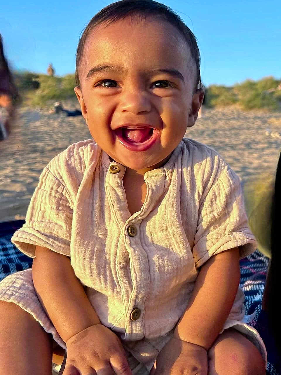 Kenzo is registered to the contest to win money with this photo: toddler, child, smiling, happy, outdoor, beach, sand, sunlight, buttoned_shirt, sitting, blanket, portrait, face, young_child, cute, natural_light, casual_clothing, daytime, skin, baby