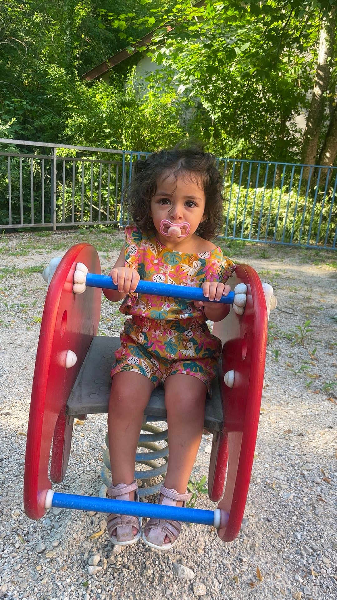 Léyna a rejoint le concours — aidez-le/la à gagner de superbes lots ! toddler, child, pacifier, playground, spring_rider, outdoor, gravel, fence, greenery, curly_hair, sandal, colorful_clothing, toy, summer, nature, daylight, young_child, sitting, play, park