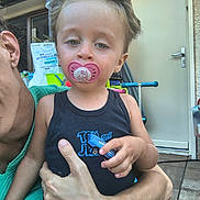 Kahil participe au concours pour gagner de l'argent avec cette photo : toddler, child, pacifier, toy_car, adult, hand, black_tank_top, green_clothing, door, scooter, outdoor, pavement, relaxed, hair, face, skin, person, holding, daylight, casual