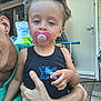 toddler, child, pacifier, toy_car, adult, hand, black_tank_top, green_clothing, door, scooter, outdoor, pavement, relaxed, hair, face, skin, person, holding, daylight, casual