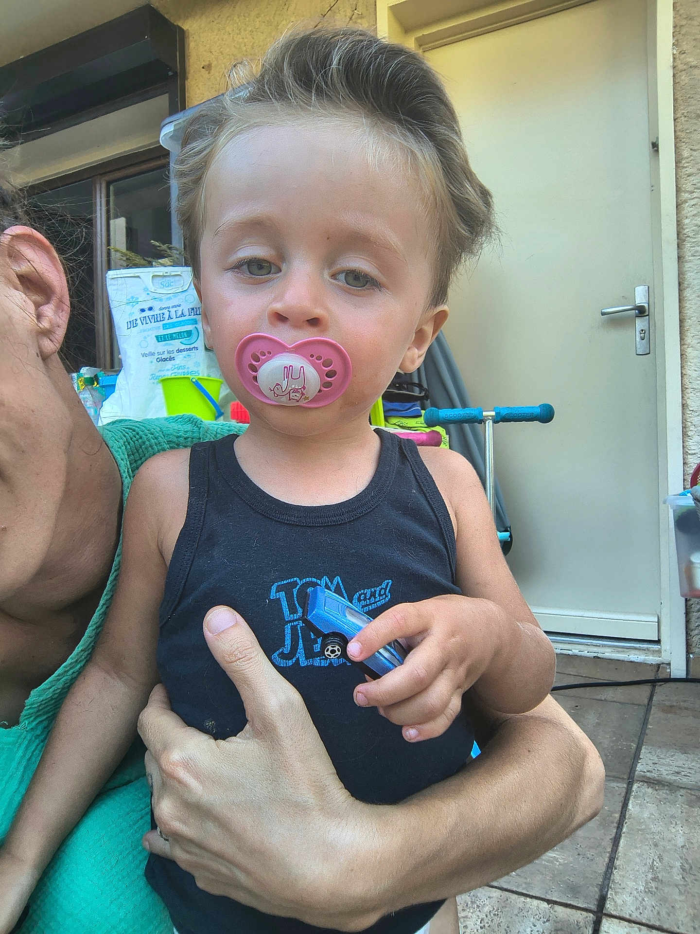 Kahil participe au concours pour gagner de l'argent avec cette photo : toddler, child, pacifier, toy_car, adult, hand, black_tank_top, green_clothing, door, scooter, outdoor, pavement, relaxed, hair, face, skin, person, holding, daylight, casual