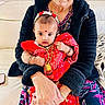 baby, child, adult, person, red_clothing, traditional_clothing, jewelry, headband, bindi, bracelet, holding, couch, black_jacket, flower, indoor, portrait, closeup, skin, hand, sitting