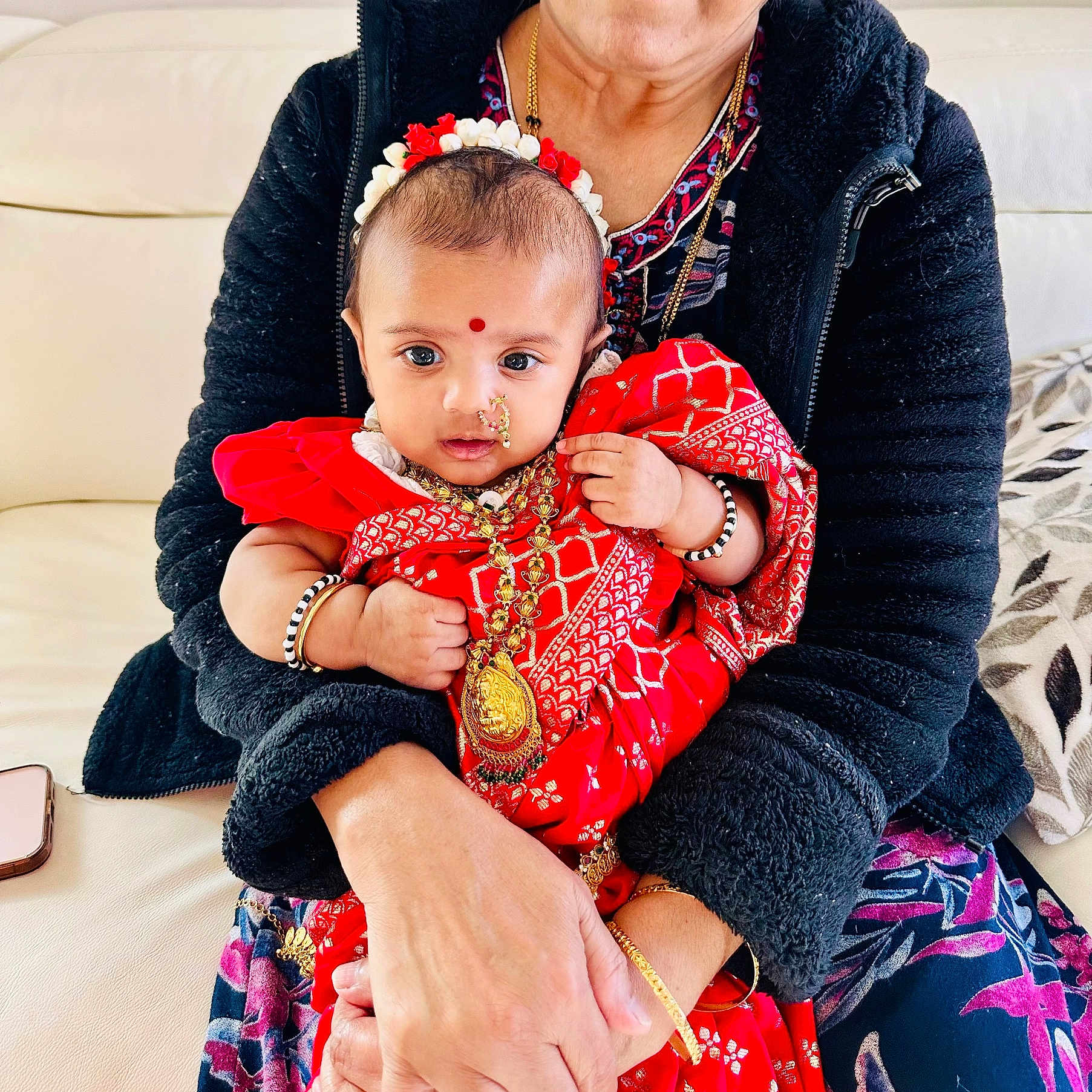 Aryahi joined the competition — help win amazing prizes! baby, child, adult, person, red_clothing, traditional_clothing, jewelry, headband, bindi, bracelet, holding, couch, black_jacket, flower, indoor, portrait, closeup, skin, hand, sitting