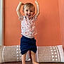 barefoot, casual_clothing, child, cushions, cute, happy, home, indoor, orange_wall, person, playful, portrait, raised_arms, shirt, shorts, smiling, standing, toddler, wooden_floor, young_child