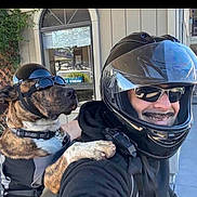 Max is registered to the contest to win money with this photo: dog, person, helmet, goggles, motorcycle, smile, outdoor, brindle, black_clothing, pet, animal, transportation, glasses, happy, casual, vehicle, friendship, companionship, protective_gear, adventure