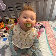 Meka is registered to the contest to win money with this photo: baby, pacifier, crib, toy, quilt, baby_clothing, bib, indoor, child, bed, cute, infant, face, hair, playroom, smile, person, colorful, plush_toy, sleepwear