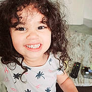 Sofia a rejoint le concours — aidez-le/la à gagner de superbes lots ! child, smiling, curly_hair, indoor, living_room, remote_control, table, shirt, patterned_clothing, happy, portrait, person, face, eyes, teeth, candid, cute, playful, young_child, toddler