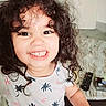 child, smiling, curly_hair, indoor, living_room, remote_control, table, shirt, patterned_clothing, happy, portrait, person, face, eyes, teeth, candid, cute, playful, young_child, toddler