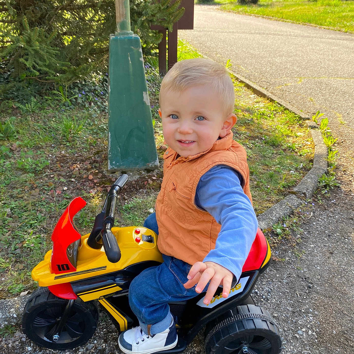 Jules a rejoint le concours — aidez-le/la à gagner de superbes lots ! blue_eyes, blue_jeans, casual_clothing, child, cute, daylight, grass, greenery, happy, orange_vest, outdoor, park, pavement, play, reaching_hand, small_vehicle, smiling, toddler, toy_motorcycle, white_shoes