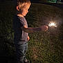 child, boy, sparkler, night, grass, outdoor, light, illumination, casual_clothing, jeans, short_sleeve, shoes, side_view, young, holding, firework, celebration, dark, portrait, curiosity