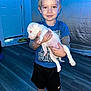 child, boy, puppy, dog, indoor, barefoot, blue_shirt, black_shorts, smile, pet, wooden_floor, door, home, person, animal, young, holding, cute, light, floor