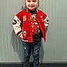 child, smiling, standing, red_jacket, jeans, sneakers, cartoon_tshirt, hair_buns, hair_accessories, indoor, confident_pose, wall, carpet, casual_clothing, happy, portrait, person, fashion, cute, young