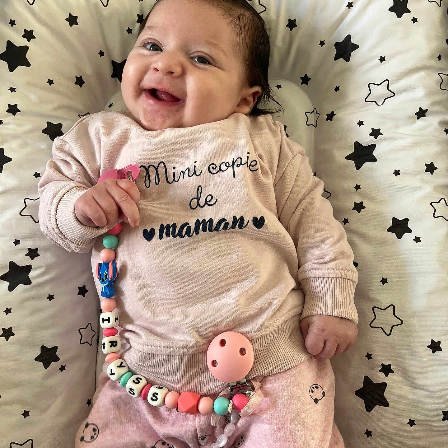 Iryss participe au concours pour gagner de l'argent avec cette photo : adorable, baby, child, comfort, cushion, cute, expression, face, hand, happy, indoors, infant, lying_down, pacifier_chain, pajamas, pink_clothing, portrait, smiling, soft_toys, star_pattern