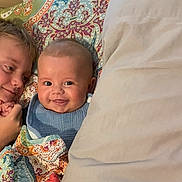 Grayson is registered to the contest to win money with this photo: baby, child, smile, bed, blanket, patterned_blanket, pillow, indoor, happy, face, person, cute, sibling, family, cozy, head, lying_down, comfort, young_child, expression