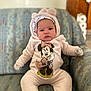 baby, child, onesie, minnie_mouse, armchair, indoor, face, cute, cozy, clothing, infant, person, seated, patterned_fabric, headwear, gloves, expression, portrait, soft_texture, home