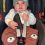 baby, child, car_seat, beanie, hat, clothing, pants, bear_design, socks, straps, seat_belt, infant, cute, indoor, person, face, hands, footwear, expression, safety