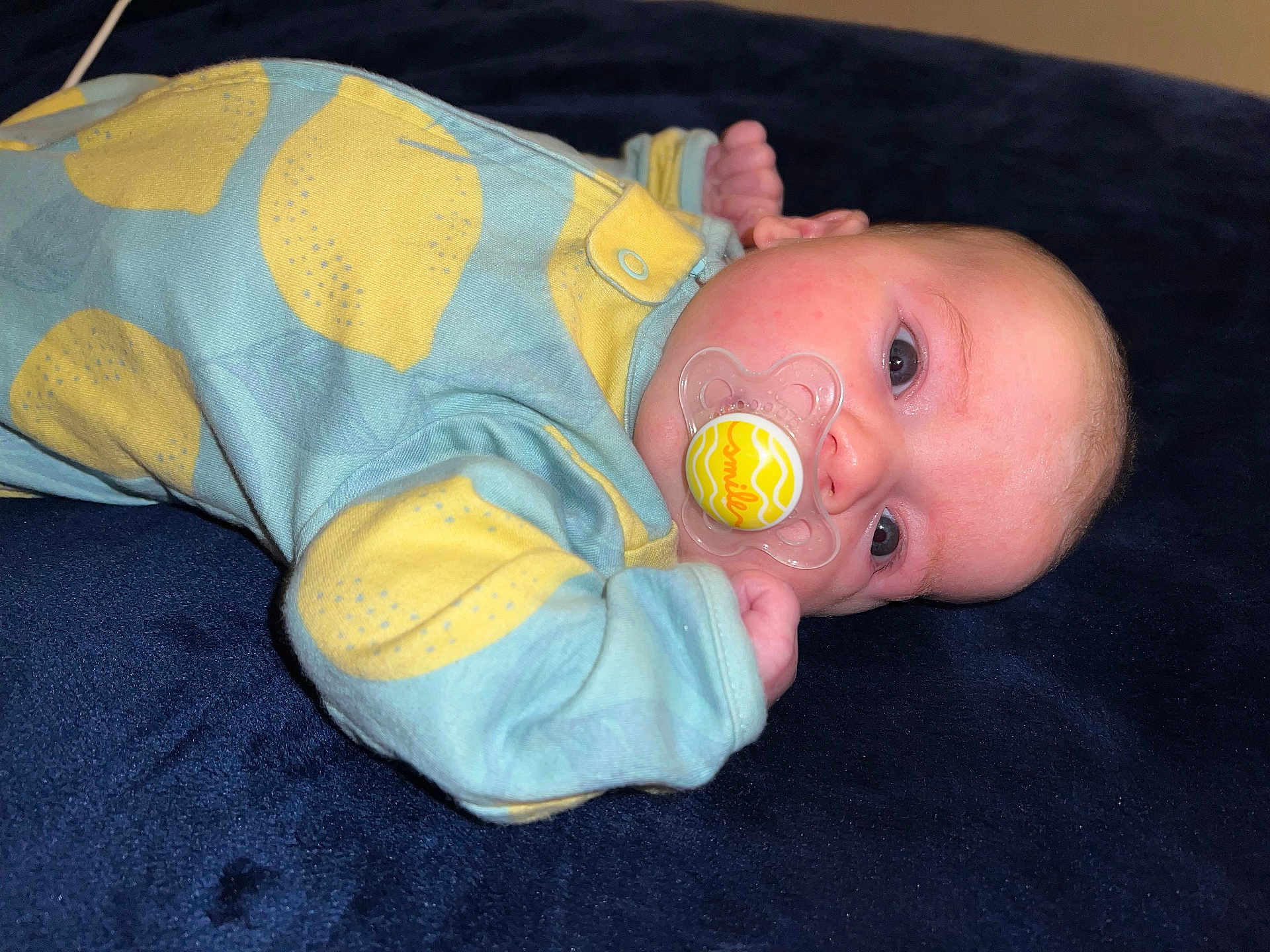 Christina is registered to the contest to win money with this photo: baby, infant, pacifier, onesie, lemon_pattern, blue_clothing, yellow, child, sleepwear, lying_down, face, skin, hand, cute, indoor, soft_lighting, expression, newborn, comfort, resting