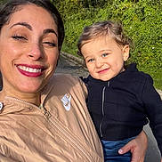 émir a rejoint le concours — aidez-le/la à gagner de superbes lots ! woman, child, toddler, smiling, winking, outdoor, daylight, greenery, curly_hair, blue_sky, jacket, zipper, holding, person, nature, casual_clothing, happy, portrait, family, road