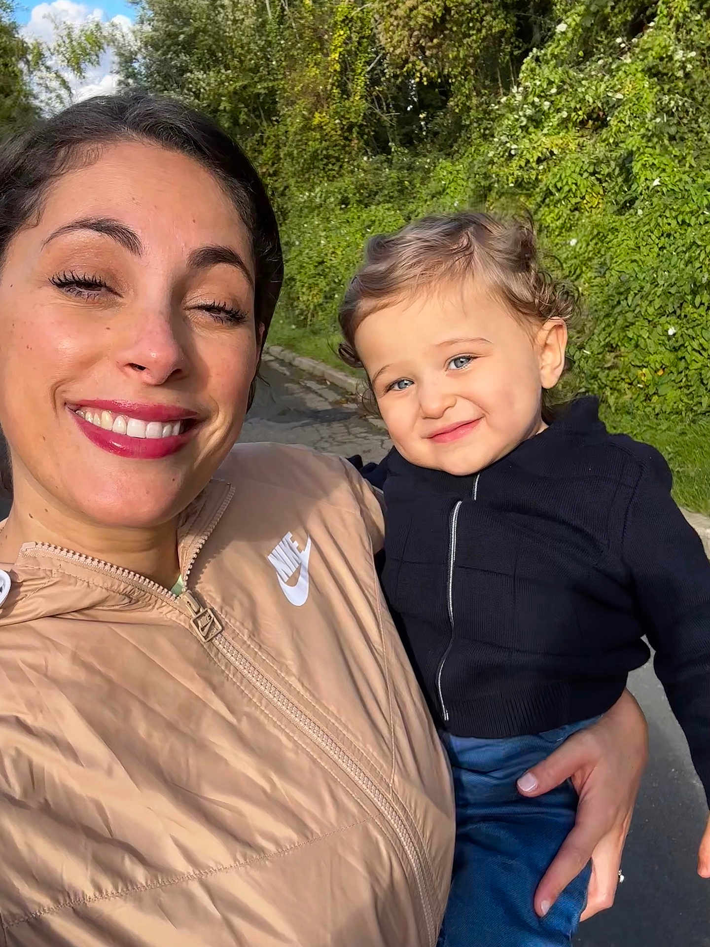 émir a rejoint le concours — aidez-le/la à gagner de superbes lots ! woman, child, toddler, smiling, winking, outdoor, daylight, greenery, curly_hair, blue_sky, jacket, zipper, holding, person, nature, casual_clothing, happy, portrait, family, road