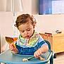 émir a rejoint le concours — aidez-le/la à gagner de superbes lots ! toddler, child, curly_hair, high_chair, denim_overalls, bib, spoon, food, fruit, indoor, lamp, wooden_furniture, table, floor, concentration, baby_food, cute, casual_clothing, person, home