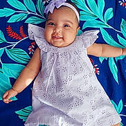 Nathaniya is registered to the contest to win money with this photo: baby, infant, smiling, dress, headband, bow, lying_down, blanket, floral_pattern, happy, cute, chubby_cheeks, person, portrait, child, playful, indoors, colorful, fabric, soft