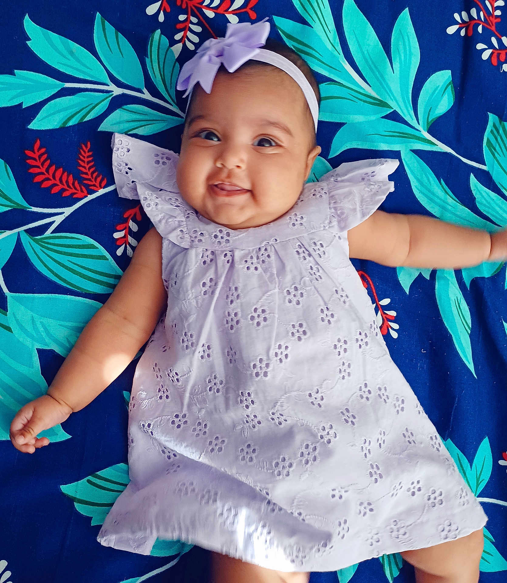 Nathaniya is registered to the contest to win money with this photo: baby, infant, smiling, dress, headband, bow, lying_down, blanket, floral_pattern, happy, cute, chubby_cheeks, person, portrait, child, playful, indoors, colorful, fabric, soft