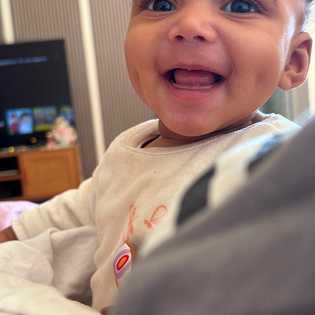 Milena a rejoint le concours — aidez-le/la à gagner de superbes lots ! baby, background_blur, blanket, child, close_up, couch, curly_hair, eyes, face, furniture, happy, indoors, onesie, portrait, smiling, soft_lighting, teeth, television, tongue, wooden_cabinet