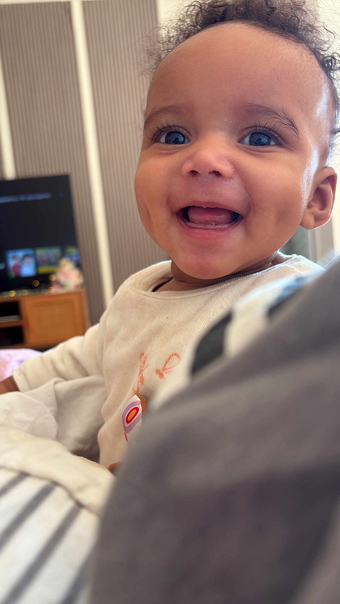 Milena a rejoint le concours — aidez-le/la à gagner de superbes lots ! baby, background_blur, blanket, child, close_up, couch, curly_hair, eyes, face, furniture, happy, indoors, onesie, portrait, smiling, soft_lighting, teeth, television, tongue, wooden_cabinet