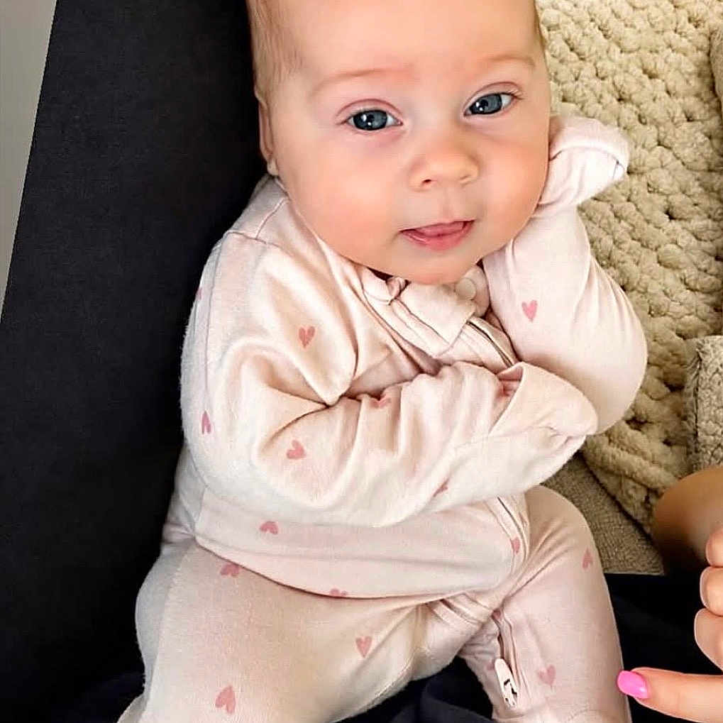 Lyla Jo is registered to the contest to win money with this photo: baby, infant, onesie, pink, hearts, face, blue_eyes, cushion, blanket, hand, nail_polish, indoor, portrait, cute, child, person, soft_lighting, cozy, seated, expression