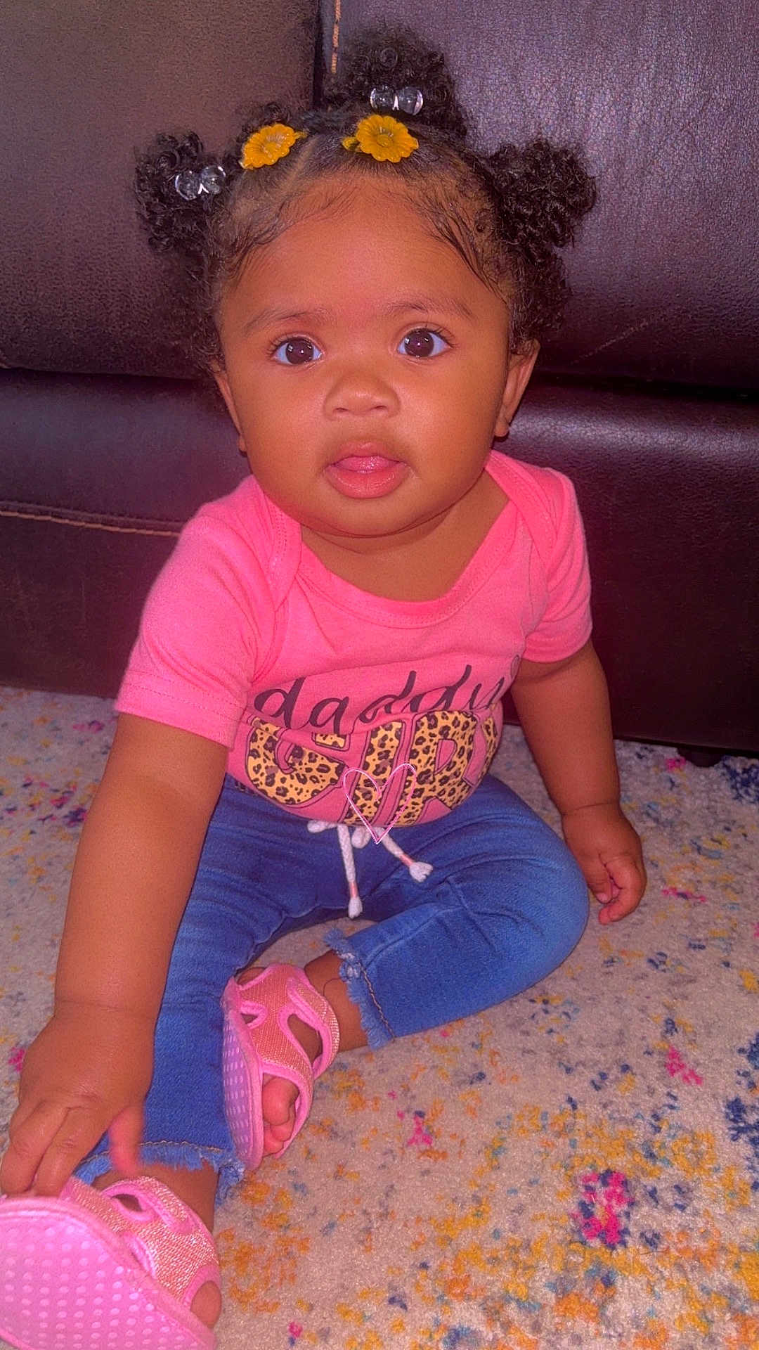Symphoni is registered to the contest to win money with this photo: baby, blue_jeans, carpet, casual_clothing, child, colorful_carpet, curious, curly_hair, cute, hair_buns, indoor, leather_couch, pink_sandals, pink_shirt, portrait, sitting, toddler, wide_eyes, yellow_flower_clips, young_child