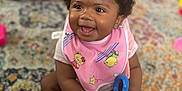 Symphoni is registered to the contest to win money with this photo: baby, bib, carpet, child, colorful, curly_hair, cute, floor, fun, happy, indoor, infant, person, pink, playful, portrait, sitting, smiling, soft_lighting, toy