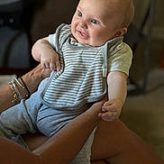 Noah joined the competition — help win amazing prizes! baby, child, smiling, indoor, person, adult_hands, bib, clothing, gray_pants, blue_eyes, short_hair, holding, sitting, happy, infant, cute, human, portrait, skin, arm